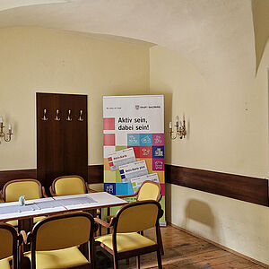 Teilansicht eines Raumes mit einem Tisch und Stühlen, links die Fensterwand, rechts der Durchgang zu anderen Räumen. Im Hintergrund ein hohes Ständerplakat der Stadt Salzburg, mit der Bewerbung der Aktiv:Karte