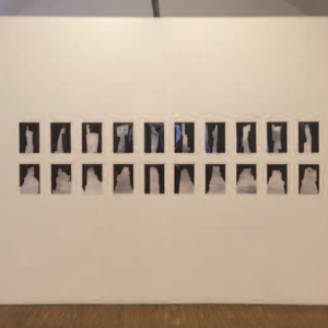 Eine weiße Wand die quer in einem Raum aufgestellt wurde; mittig auf dieser Wand sind in zwei Reihen je 10 Fotos übereinander angebracht. Links und rechts dieser Wand ist noch Platz zum vorbei gehen.