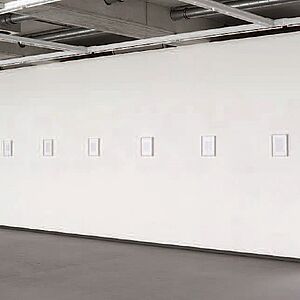 Eine weiße Wand in einem Raum mit abgehängten Deckenstreben (die Wand reicht bis zu diesen Streben) mit sieben kleinen mittig aufgehängten Bildern
