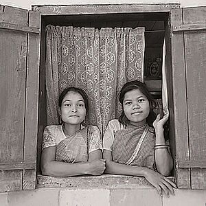 schwarz-weiß Fotografie zweier junger indischer Mädchen die aus einem Fenster mit seitlich geöffneten Fensterläden schauen