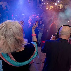 Das Bild zeigt eine Tanzveranstaltung. Im Vordergrund sieht man einen DJ von hinten. Er trägt Kopfhörer und steht an einem Mischpult mit vielen Knöpfen und Lichtern. Neben ihm steht eine Frau mit grauen Haaren. Sie hebt ihre Arme und klatscht im Takt. Viele Menschen tanzen vor der Bühne. Der Raum ist voll und in buntes Licht getaucht. Es gibt Nebel im Raum, der das Licht weich erscheinen lässt. Die Stimmung wirkt lebendig und fröhlich.