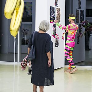 Der Künstler in einem bunten Ganzkörper Overall vor einem Bild, eine Frau im Vorderrgrund beobachtet den Künstler bei seiner Performance