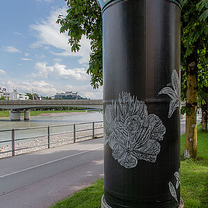 Litfaßsäule mit dem Kunstplakat, im Hintergrund der Geh- und Radweg entlang der Salzach und der Fluss 