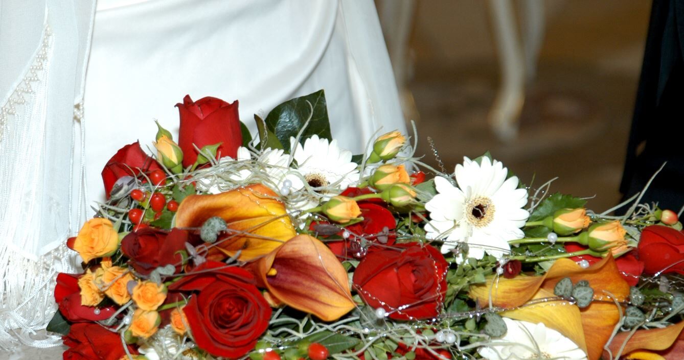 Blumenstrauss mit einem weißen Hochzeitskleid im Hintergrund.