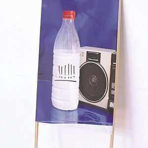 Eine Kunststoffmilchflasche steht vor einem Kompakt-Kassettenrecorder von dem nur ein Lautsprecher im Bild ist. Die Flasche ist mit mehreren Rufzeichen die unterstrichen sind angezeichnet.