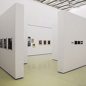 Zwei weiße Wände eines Raumes mit aufgehängten Bildern, im Vordergrund des Raumes zwei frei, im rechten Winkel zueinander stehende Wände mit aufgehängten Bildern.