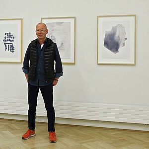 Foto von dem Künstler Josef Schwaiger, hinter ihm 3 Bilder auf einer weißen Wand