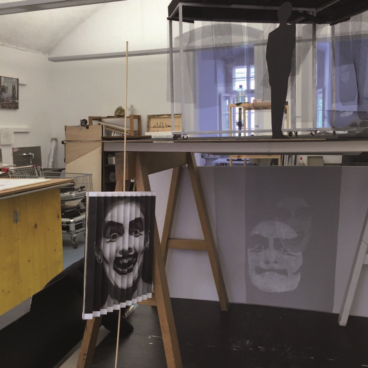 Werkstatt mit aufgebockter hoher Tischplatte auf der eine Installation steht, davor ein Bild mit einem stark geschmickten schwarz-weißem Gesicht - gefaltet wie ein Fächer - an einem senkrechten Stab. 