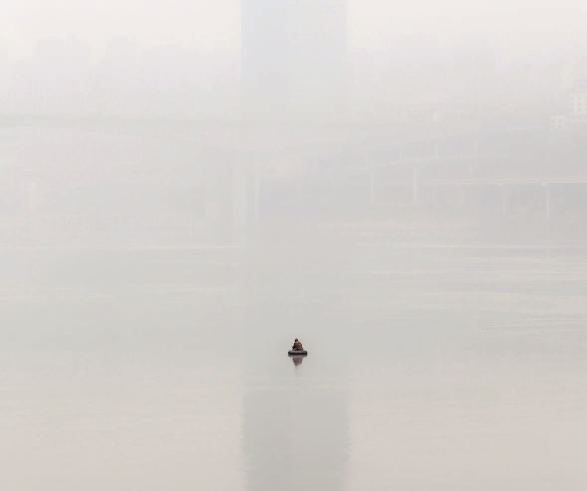 Nebelverhangenes Bild mit einer Person auf dem Wasser in einem sehr kleinen runden Schlauchboot sitzend. Im Hintergrund im Nebel kaum erkennbare Gebäudestrukturen.