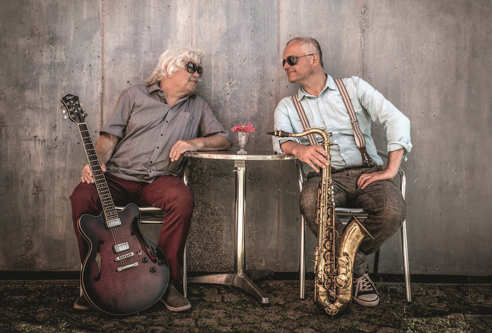 Punzenberger und Berger zwei Musiker nebeneinander an einem Bistrotisch sitzend, de linke Musiker hält eine Gitarre in der Hand der rechte Musiker ein Saxophon.